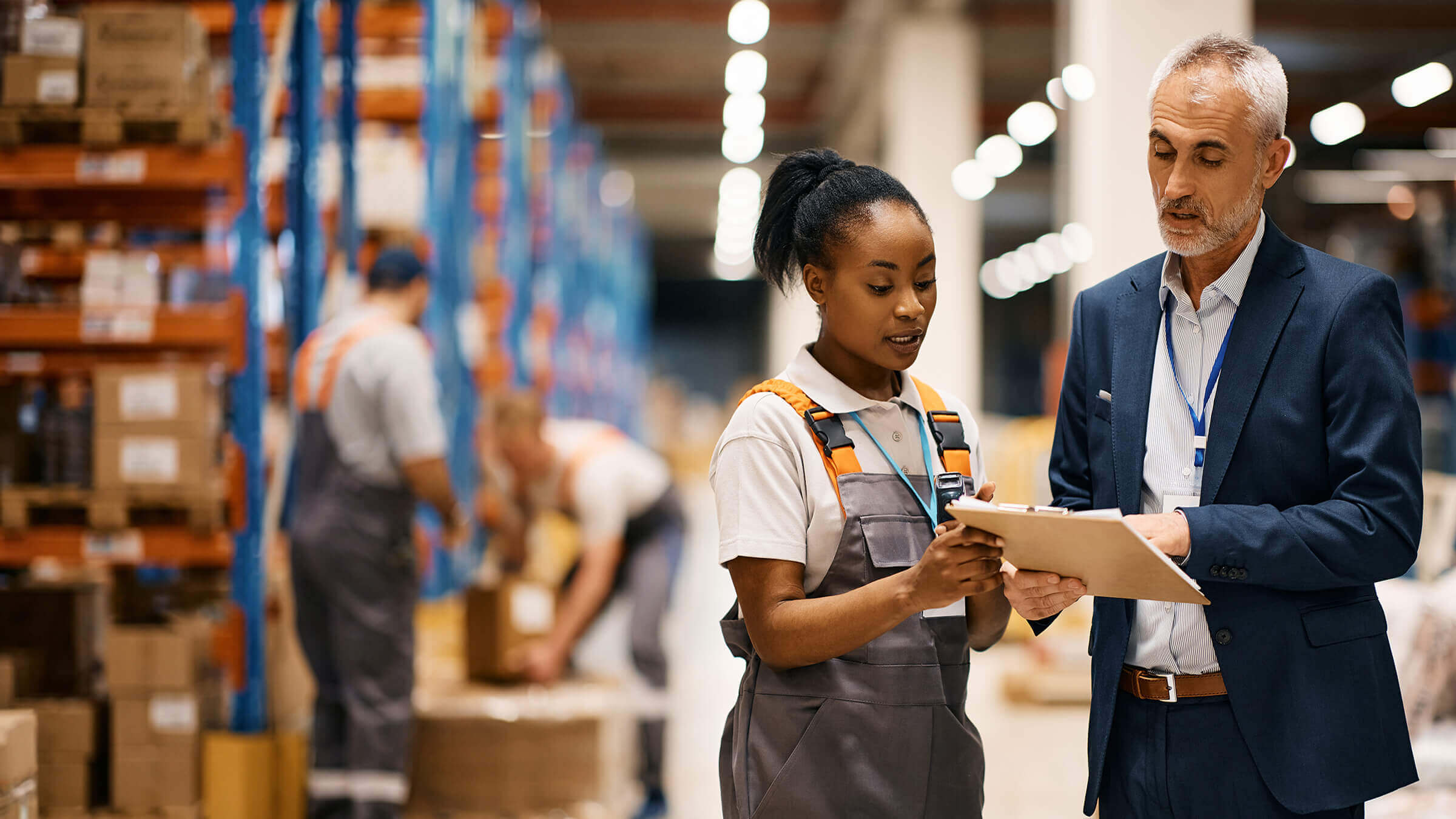 Warehouse supervisor reviewing inventory details with a logistics worker on the warehouse floor while orders are prepared in the background.