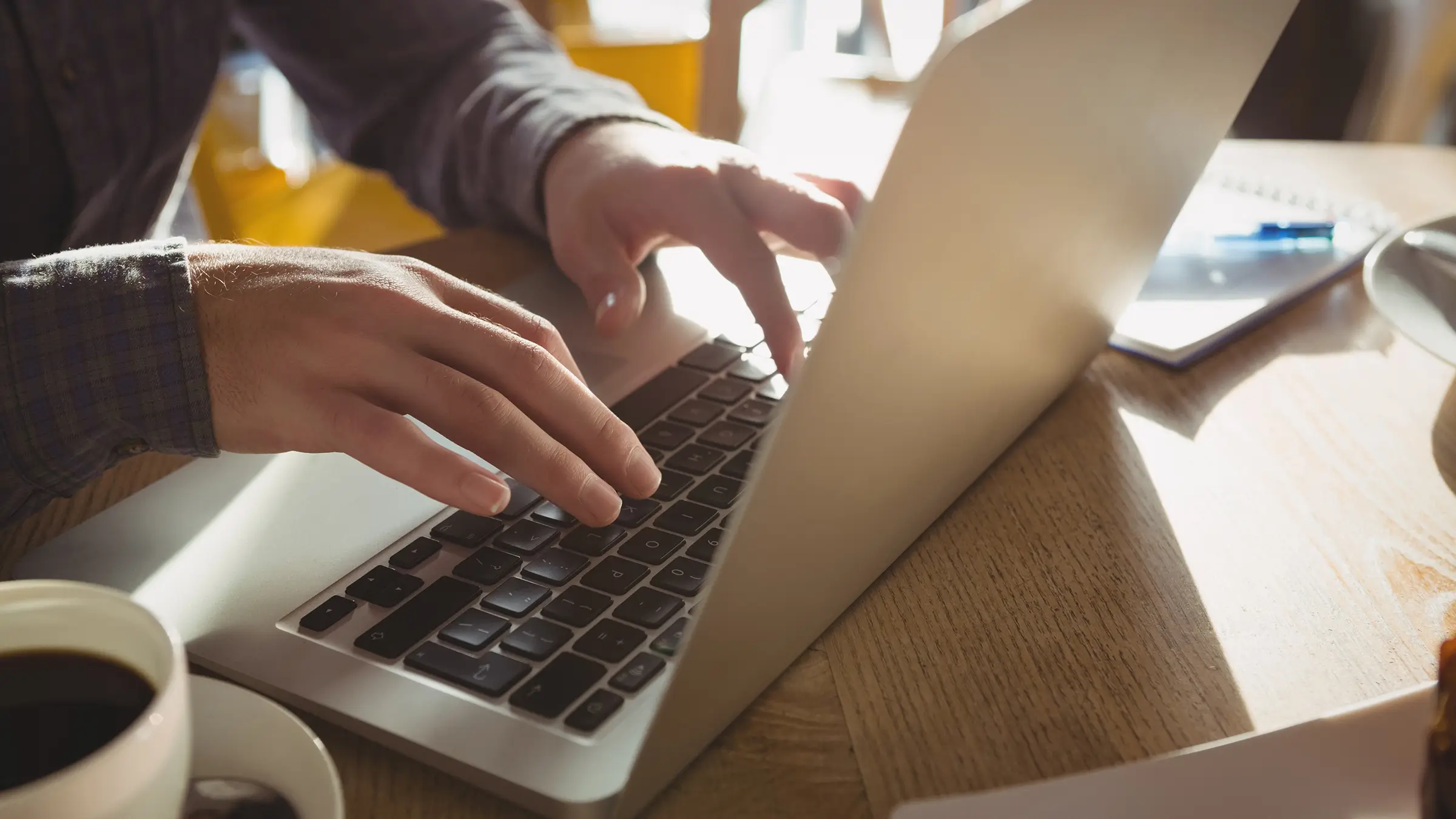 Person typing a blog on laptop at wooden desk with coffee and notebook in warm natural light