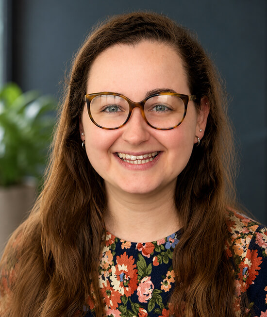 Professional headshot of a smiling woman with long brown hair wearing a floral shirt.