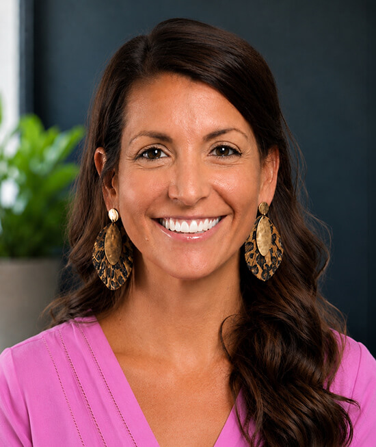 Professional headshot of a smiling woman in pink shirt with dark hair.